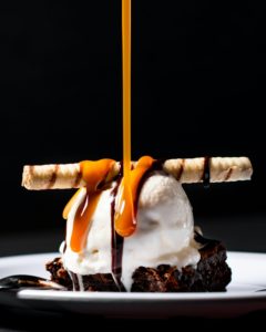 Brownie on a white plate topped by ice cream and caramel sauce being poured on it from above